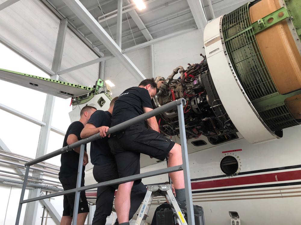 Three technicians on a work platform servicing an opened turbofan engine on an aircraft in a hangar
